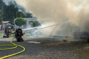 "Marie fängt Feuer - Vergeben und Vergessen": Marie (Christine Eixenberger) kniet zusammen mit einem Kollegen der Feuerwehr auf dem Boden des Campingplatzes Beide zielen mit Feuerwehrschläuchen auf brennende Wohnwagen, die sich hinter einer Rauchwolke befinden.