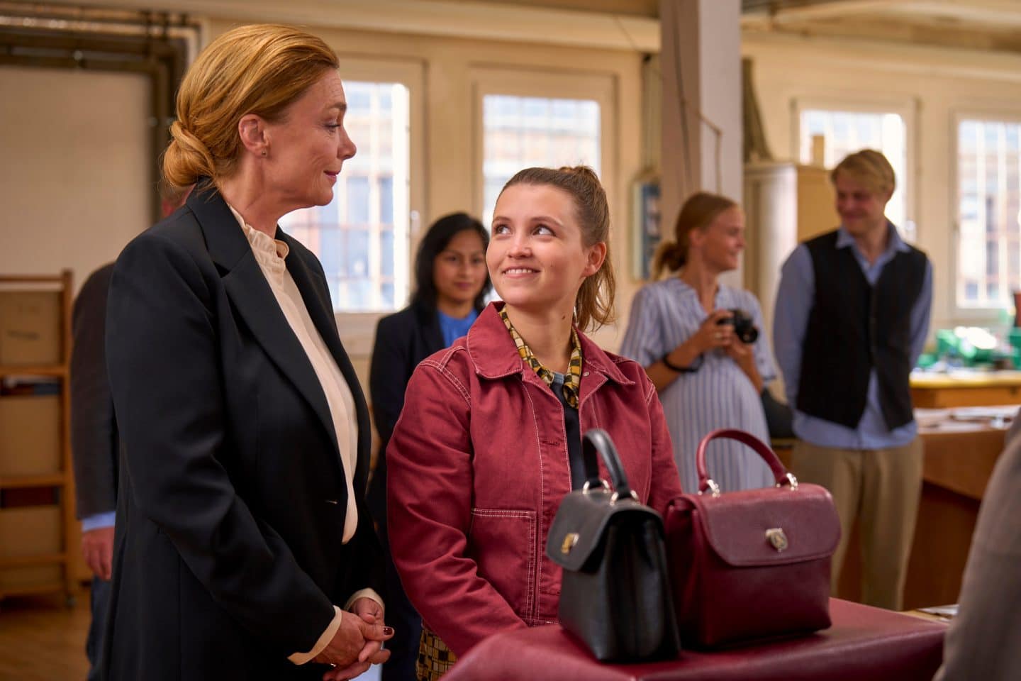 "Zimmer im Grünen - Herzenswege": Heide (Aglaia Szyszkowitz) und Lisa (Janina Fautz) stehen nebeneinander in einem großen Raum. Vor ihnen stehen zwei Modelle der Krapp-Handtaschen. Lisa schaut lächelnd zu Heide, im Hintergrund stehen weitere Mitarbeiter (Komparsen) der Firma Krapp.
