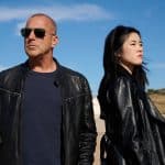 "Yvonne und der Tod": Ingo Thiel (Heino Ferch) steht auf einem sonnigen Platz und schaut mit aufgesetzter Sonnenbrille in die Ferne. Lee Sooyoung (Kotbong Yang) steht neben ihm. Sie blickt in die andere Richtung und hat ihm den Rücken zugewandt.