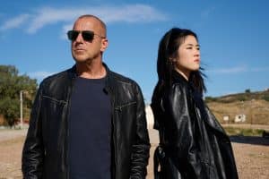 "Yvonne und der Tod": Ingo Thiel (Heino Ferch) steht auf einem sonnigen Platz und schaut mit aufgesetzter Sonnenbrille in die Ferne. Lee Sooyoung (Kotbong Yang) steht neben ihm. Sie blickt in die andere Richtung und hat ihm den Rücken zugewandt.