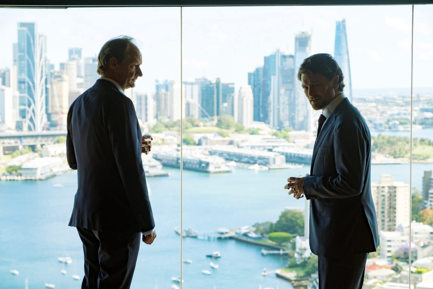 "North Shore – Tod in Sydney – Sophies Laptop": Lloyd (Rob Carlton) und Simon (Dan Spielman) stehen in einem Büro vor einer großen Fensterfront. Draußen liegt die Skyline von Sydney mit dem Hafen..
