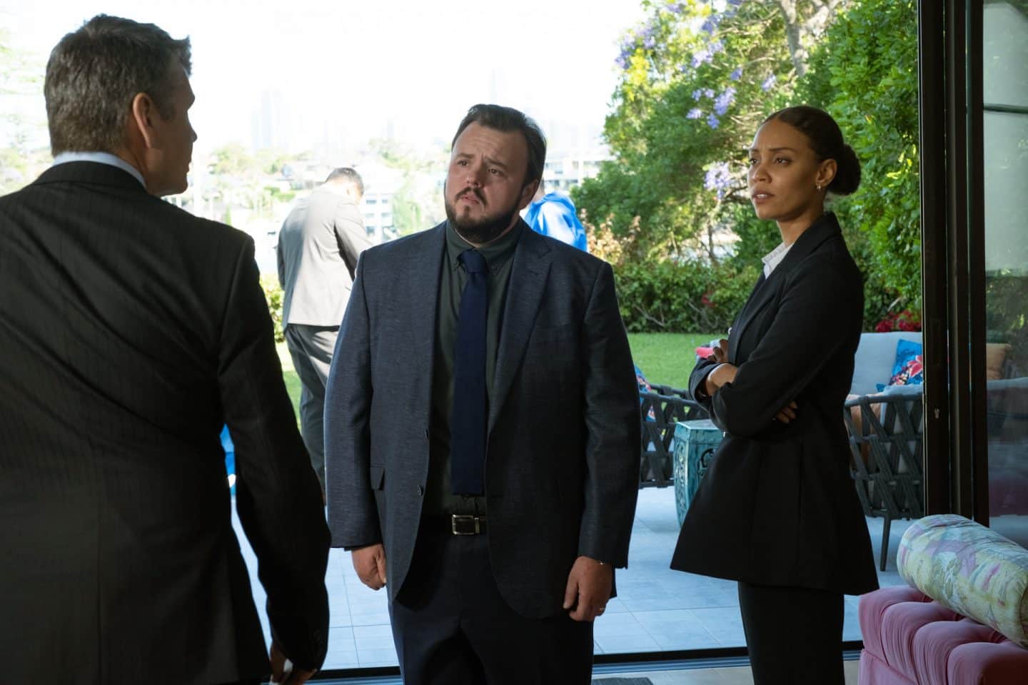 "North Shore – Tod in Sydney – Die Spur des Blutes": Meg (Kirsty Sturgess) und Max (John Bradley) stehen in einem Raum vor einer geöffneten Tür, die auf eine Terrasse führt. Ihnen gegenüber steht Anwalt Gary Weaver (Michael Beckley). Im Hintergrund ist ein weiterer Mann zu sehen.
