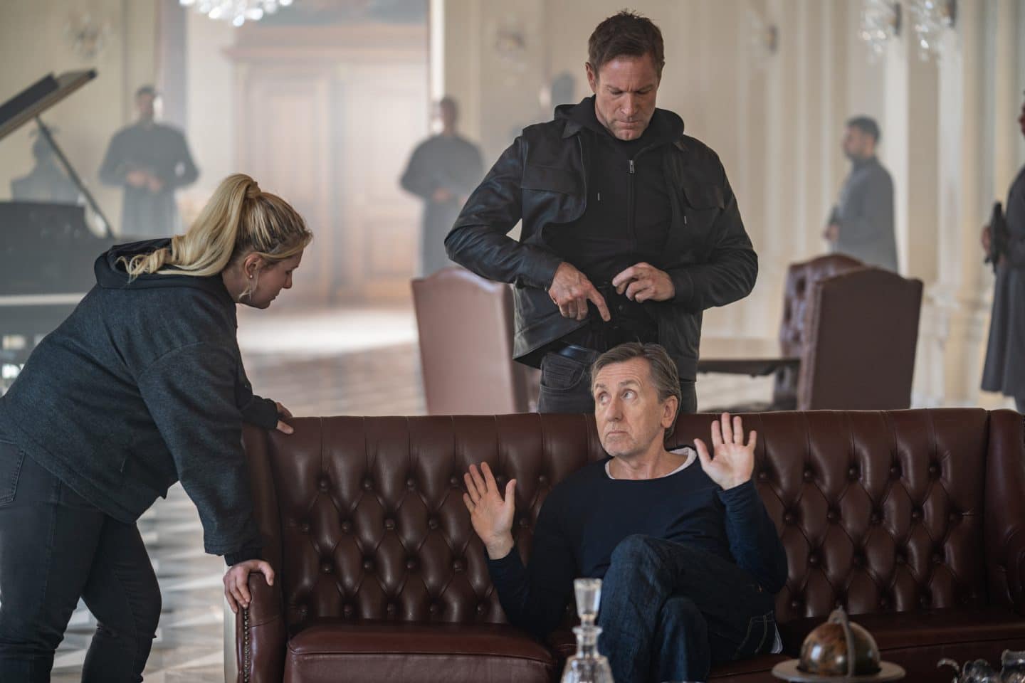 "Geheimsache Malta": Angler (Tim Roth) sitzt auf einem Ledersofa und hält die Hände in einer nichts wissenden Geste nach oben. Er sieht zur Seite. Dort steht Kacey (Abigail Breslin), die ihn ebenfalls anschaut. Hinter dem Sofa steht Shaw (Aaron Eckhart). Er scheint eine Waffe aus seiner Hose zu ziehen. Im Hintergrund sind verschwommen bewaffnete Bodyguards (Komparsen) in einem Halbkreis zu sehen.
