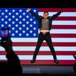 "Die Elon-Musk-Story - Superreich und supermächtig?": Elon Musk springt auf einer Bühne in die Luft, hinter ihm hängt die amerikanische Flagge.