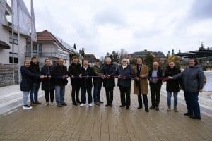 Eine Gruppe von Menschen steht in einer Reihe und hält ein Band, das für eine feierliche Eröffnung durchschnitten werden soll. Die Personen sind in warme Kleidung gehüllt, was auf kühles Wetter hindeutet. Im Hintergrund sind Gebäude mit traditioneller Architektur zu sehen, darunter ein Gebäude mit gelber Fassade und grünen Türmen sowie eine Pension mit Solarpanelen auf dem Dach. Rechts im Bild stehen Stehtische mit Getränken, was auf eine offizielle Veranstaltung hinweist.