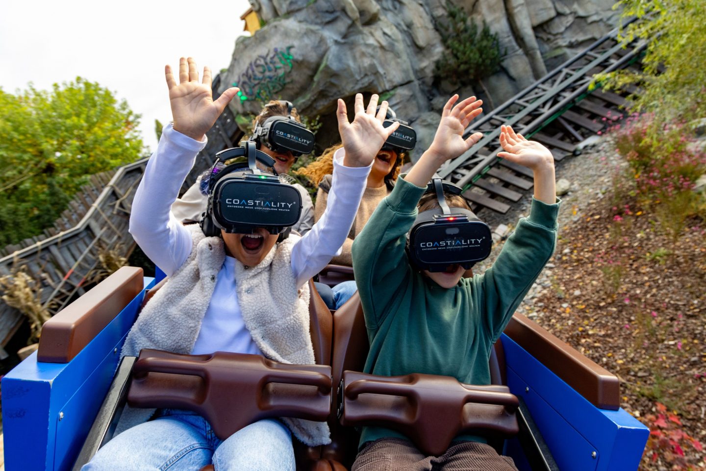 Zwei begeisterte Kinder mit VR-Brillen erleben eine Achterbahnfahrt im Europa-Park. Mit erhobenen Armen genießen sie das virtuelle Coastiality-Erlebnis, während die Schienen und die Felslandschaft im Hintergrund die immersive Kombination aus realer und digitaler Welt unterstreichen.