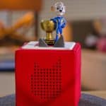 Ein rotes Toniebox-Hörspielgerät steht auf einem runden Filzuntersetzer auf einem Holztisch. Auf der Box befindet sich die Tonie-Figur Ed Euromaus, bekannt aus dem Europa-Park, in einem blauen Rennanzug, die eine goldene Trophäe hält. Im Hintergrund ist ein Wohnzimmer mit einer Lampe und verschwommenen Spielzeugfiguren zu sehen.