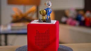 Ein rotes Toniebox-Hörspielgerät steht auf einem runden Filzuntersetzer auf einem Holztisch. Auf der Box befindet sich die Tonie-Figur Ed Euromaus, bekannt aus dem Europa-Park, in einem blauen Rennanzug, die eine goldene Trophäe hält. Im Hintergrund ist ein Wohnzimmer mit einer Lampe und verschwommenen Spielzeugfiguren zu sehen.