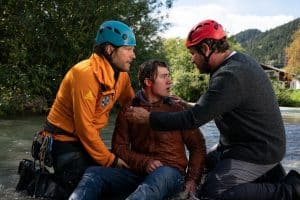 "Die Bergretter: Im Gipfelbuch": Bergretter Markus (Sebastian Ströbel) kniet im Wasser neben Felix Hertinger (Alessandro Schuster), der von seinem Vater Jonas (Stephan Szász) an den Schultern gehalten wird. Markus und Jonas tragen Helme, Felix sitzt durchnässt und aus dem Ohr blutend im Wasser zwischen den beiden.