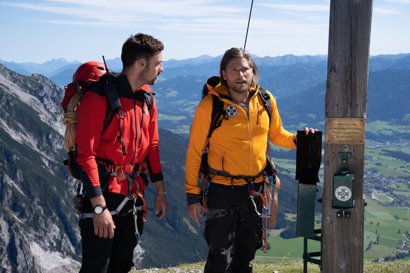 "Die Bergretter: Im Gipfelbuch": Die Bergretter Markus (Sebastian Ströbel) und Simon (Ferdinand Seebacher) stehen am Gipfel eines Berges und wirken überrascht. Markus hält mit einer Hand eine Klappe fest, die normalerweise das Gipfelbuch sichert.
