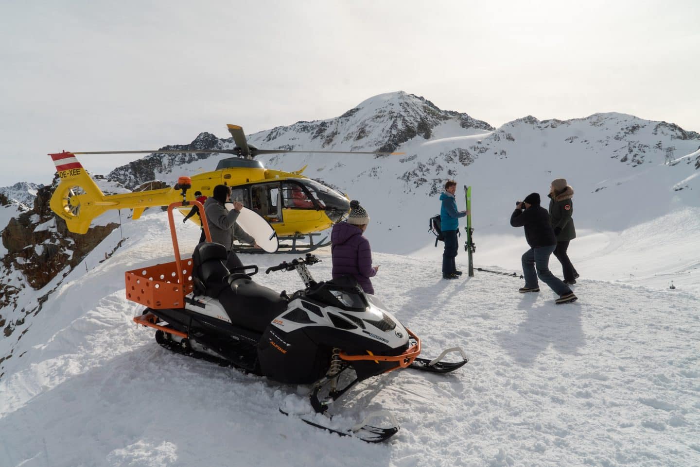"Die Bergretter: Eiskalte Wahrheit (1)": Lisa Kronach (Lena Meckel) steht an ein Schneemobil gelehnt und blickt zu Daniel Faltermayer (Julian Felix), der von zwei Fotografen vor einem Helikopter abgelichtet wird.
