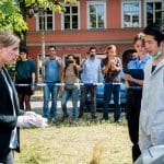 "SOKO Potsdam - Du bist schuld": Sophie Pohlmann (Katrin Jaehne) steht mit Thomas Brandner (Yung Ngo) auf einer Wiese. Thomas Brandner trägt einen weißen Schutzanzug und einen Mundschutz, den er nach unten gezogen hat. Sophie trägt weiße Gummihandschuhe. Die beiden blicken sich ernst an. Neben Thomas steht eine Polizistin (Komparsin) mit einem Klemmbrett in der Hand. Im Hintergrund stehen Passanten (Komparsen) hinter einem Absperrband und machen Fotos vom Tatort. Hinter ihnen sieht man ein Auto und ein Gebäude mit der Aufschrift "Sanitätshaus am Schloss". Die Sonne scheint.