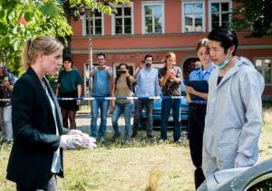 "SOKO Potsdam - Du bist schuld": Sophie Pohlmann (Katrin Jaehne) steht mit Thomas Brandner (Yung Ngo) auf einer Wiese. Thomas Brandner trägt einen weißen Schutzanzug und einen Mundschutz, den er nach unten gezogen hat. Sophie trägt weiße Gummihandschuhe. Die beiden blicken sich ernst an. Neben Thomas steht eine Polizistin (Komparsin) mit einem Klemmbrett in der Hand. Im Hintergrund stehen Passanten (Komparsen) hinter einem Absperrband und machen Fotos vom Tatort. Hinter ihnen sieht man ein Auto und ein Gebäude mit der Aufschrift "Sanitätshaus am Schloss". Die Sonne scheint.
