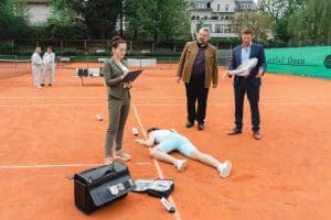 "Die Rosenheim-Cops - Alles hat ein Ende": Pathologin Atay (Sevda Polat), Kommissar Stadler (Dieter Fischer) und Kommissar Hansen (Igor Jeftić) stehen um das Mordopfer herum, das bäuchlings auf dem Tennisplatz liegt. Kommissar Hansen hält einen in transparente Folie verpackten Tennisschläger in der Hand.