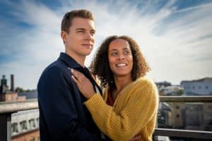 "Inga Lindström: Geliebter Feind": Elli (Pia Amofa-Antwi) und Lucas (Gerrit Klein) stehen aneinander gelehnt auf der Dachterrasse der "Berginvest"-Niederlassung in Stockholm. Im Hintergrund strahlend blauer Himmel mit Wolken, Elli hat ihre linke Hand auf Lucas' Brust gelegt. Beide schauen rechts an der Kamera vorbei in die Ferne.