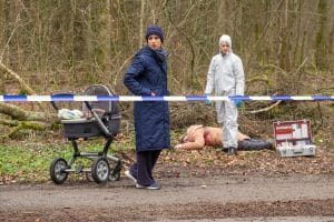 "Der Alte - Zwischenstation": Annabell Lorenz (Stephanie Stumph) steht auf einem Waldweg, hinter einem Absperrband an einem Kinderwagen und schaut sich um. Im Hintergrund liegt die Tote Marlies Thalmaier (Isabella Surel). Dr. Luisa Geiger (Sidonie von Krosigk) steht neben der Leiche und sieht Richtung Annabell Lorenz.
