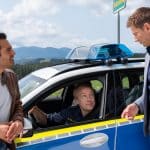 "Die Rosenheim-Cops - Hansens neuer Bruder": Kommissar Kaya (Baran Hêvî) und Kommissar Hansen (Igor Jeftić) lehnen am Polizeifahrzeug von Polizeihauptmeister Mohr (Max Müller), der am Steuer sitzt und Hansen lächelnd ansieht.