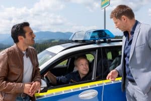"Die Rosenheim-Cops - Hansens neuer Bruder": Kommissar Kaya (Baran Hêvî) und Kommissar Hansen (Igor Jeftić) lehnen am Polizeifahrzeug von Polizeihauptmeister Mohr (Max Müller), der am Steuer sitzt und Hansen lächelnd ansieht.