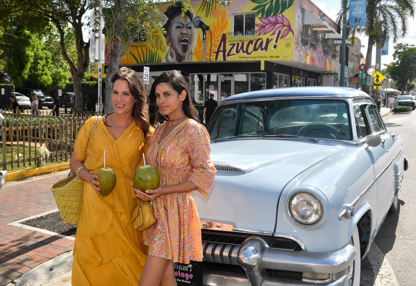 "Das Traumschiff: Miami": Im kubanischen Viertel: Dr. Jessica Delgado (Collien Ulmen-Fernandes) steht neben ihrer Freundin Maike Kappes (Birthe Wolter) vor einem Oldtimer. Sie halten Trinkkokosnüsse in den Händen und blicken lächelnd in die Ferne. Im Hintergrund ist ein Gebäude bunt bemalt.
