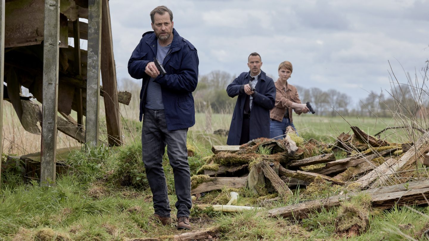 "Ostfriesenfluch": Kommissar Frank Weller (Christian Erdmann) pirscht sich mit der Waffe in der Hand auf unwegsamen verlassenem Gelände voran. Seine Kollegen Rupert (Barnaby Metschurat) und Larissa Jusewicz (Maria Magdalena Wardzinska) stehen etwas hinter ihm, haben ebenfalls jeweils eine Waffe in der Hand und sichern.
