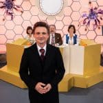 "Rosenthal": 70er Jahre: Hans Rosenthal steht im schwarzen Anzug vor einer Wabenwand im Studio. Hinter ihm sitzt seine Ratejury, Mady Riehl (Theresia Wald), "Ekki" Fritsch (Timo Dierkes) und Brigitte Xander (Katharina Völkl), an Pulten.