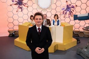 "Rosenthal": 70er Jahre: Hans Rosenthal steht im schwarzen Anzug vor einer Wabenwand im Studio. Hinter ihm sitzt seine Ratejury, Mady Riehl (Theresia Wald), "Ekki" Fritsch (Timo Dierkes) und Brigitte Xander (Katharina Völkl), an Pulten.