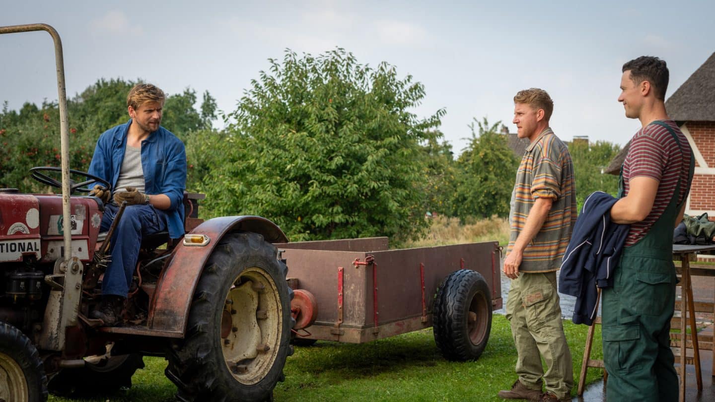 "Neuer Wind im Alten Land - Erntezeit": Paul Harms (Steve Windolf) sitzt auf einem roten alten Traktor mit Anhänger. Marek (Klaudiusz Kaufmann) und Kasimir Podolski (Mateusz Grabowsk) stehen Paul zur Begrüßung gegenüber.
