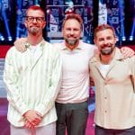 Joko Winterscheidt; Steven Gätjen; Klaas Heufer-Umlauf stehen Arm in Arm auf einer bunt beleuchteten TV-Showbühne mit dem Logo „Joko & Klaas gegen ProSieben“ im Hintergrund. Links trägt einer ein gestreiftes Hemd, in der Mitte ein Mann mit weißem Pullover, rechts ein Mann in beigefarbener Jacke. Alle drei lächeln in die Kamera.
