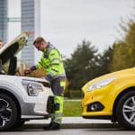 Ein ADAC-Pannenhelfer unterstützt eine Frau bei einer Autopanne, indem er unter die geöffnete Motorhaube eines weißen Fahrzeugs schaut. Im Vordergrund steht ein gelbes ADAC-Pannenhilfefahrzeug. Im Hintergrund sind Hochhäuser und Bäume zu sehen.