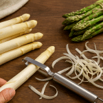 Frischer weißer und grüner Spargel auf einem rustikalen Holztisch, eine Hand schält eine Spargelstange, daneben liegt ein Bund grüner Spargel mit Küchengarn – natürliches Licht betont die frischen Farben und Texturen.
