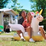 Sascha Bohner und Raffaela sitzen gemeinsam auf einem aufblasbaren Einhorn vor ihrem Wohnwagen auf dem Campingplatz Marina di Venezia – strahlend bei Sonnenschein, umgeben von Campingdeko und grüner Natur.
