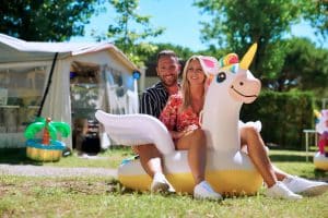 Sascha Bohner und Raffaela sitzen gemeinsam auf einem aufblasbaren Einhorn vor ihrem Wohnwagen auf dem Campingplatz Marina di Venezia – strahlend bei Sonnenschein, umgeben von Campingdeko und grüner Natur.