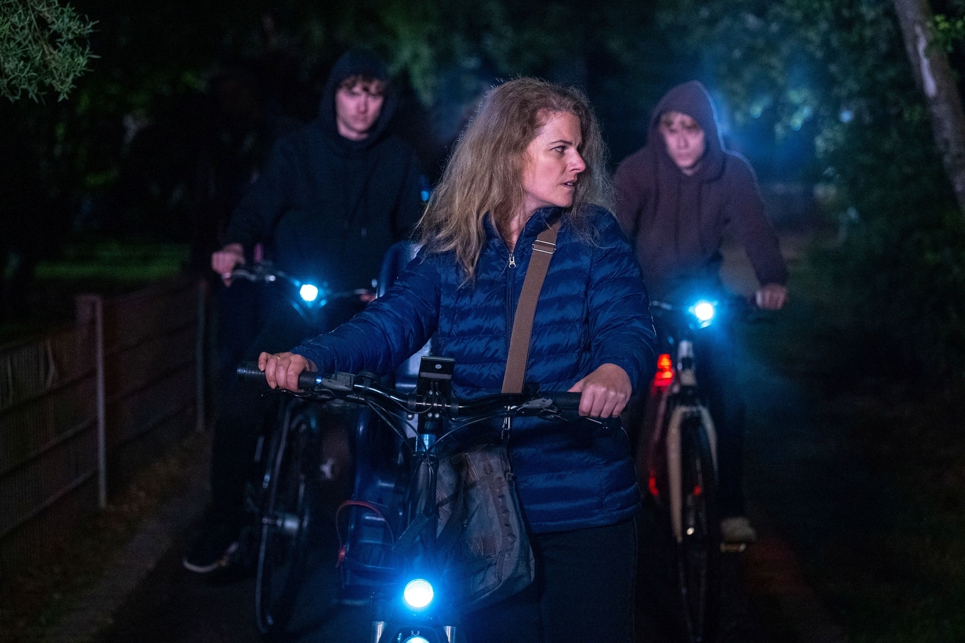 "Aktenzeichen XY... Ungelöst": Nachgestellte Szene: Eine Frau schiebt bei Nacht ein Fahrrad und schaut ängstlich halb zur Seite. Dahinter folgen zwei weitere Personen in dunkler Kleidung mit Kapuzen ebenfalls mit Fahrrädern. Alle Fahrräder haben eingeschaltete Frontlichter, die den Weg vor ihnen beleuchten. Im Hintergrund sind Bäume und Pflanzen in der Dunkelheit erkennbar.