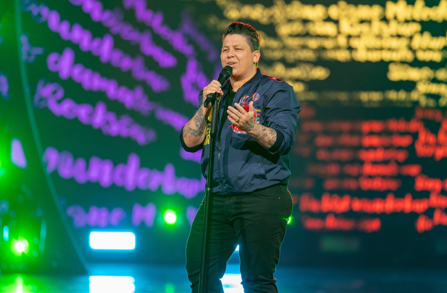 "Die Giovanni Zarrella Show": Kerstin Ott steht auf der Bühne und singt in ein Standmikrofon. Sie trägt eine dunkelblaue Bomberjacke mit Patches sowie eine dunkle Hose. Im Hintergrund ist eine farbenfrohe LED-Wand mit unscharfen Schriftzügen in lila, gelb und rot zu sehen.
