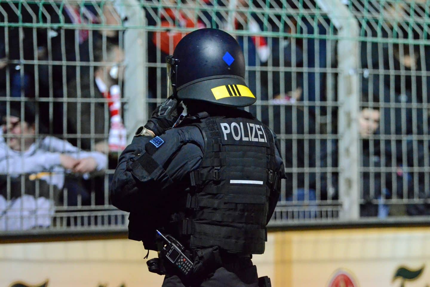 Polizist in Schutzkleidung mit Helm und Funkgerät überwacht den Zuschauerbereich in einem Fußballstadion.
