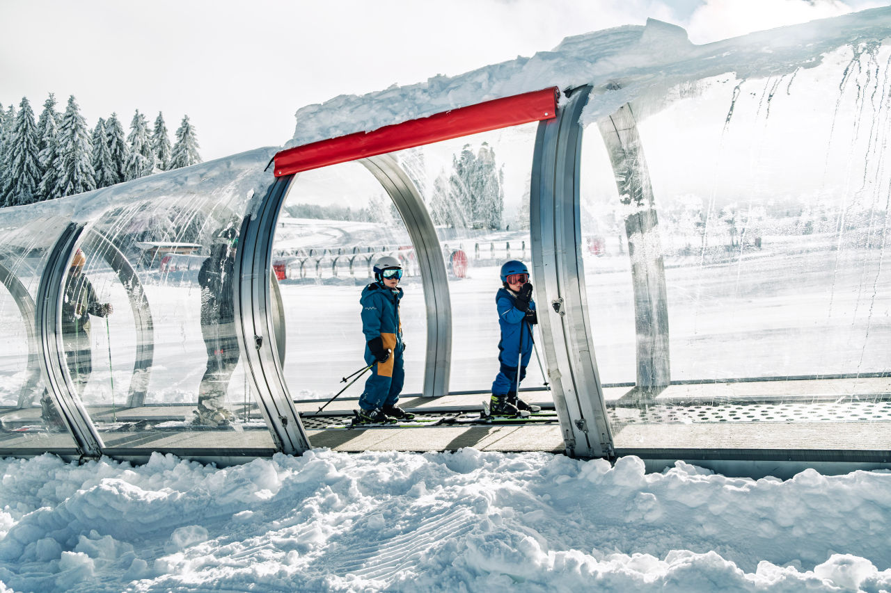 Hier ist Skifahren noch bezahlbar: Das günstigste Skigebiet im DACH-Raum liegt in Baden-Württemberg 1 Eine Familie liftet auf dem Förderband mit Überdachung.