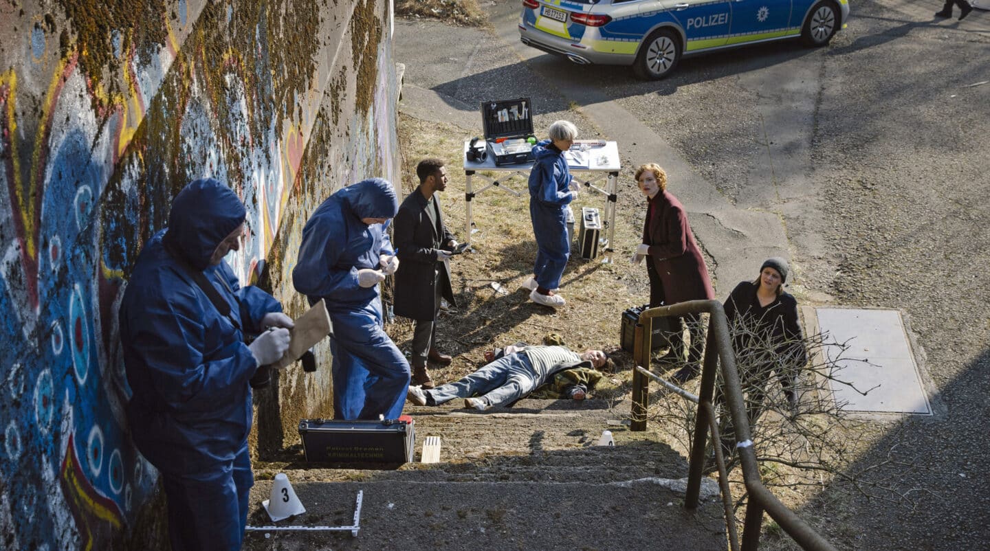 Kriminaltechniker untersuchen einen Tatort mit einem leblosen Körper und Polizeiauto im Hintergrund.