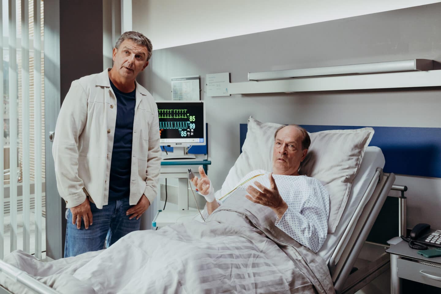 "Der Bergdoktor: Wünsche": In der Klinik: Michael Fellner (Herbert Knaup) liegt in einem Krankenbett, er trägt ein Patientenhemd und ist an einen Überwachungsmonitor angeschlossen. Er blickt gestikulierend zur Seite. Neben seinem Bett steht Dr. Martin Gruber (Hans Sigl) und blickt ebenfalls in diese Richtung. Er wirkt überrascht.