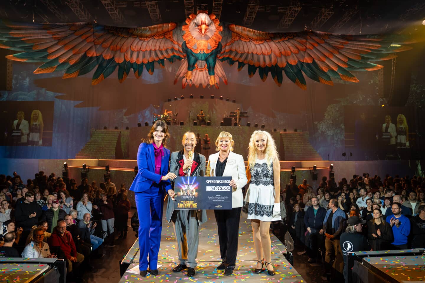 Miriam Mack, DJ BoBo, Mauritia Mack und Nancy Baumann stehen auf der Bühne der Europa-Park Arena und präsentieren einen Spendenscheck vor Publikum während der Tourpremiere.
