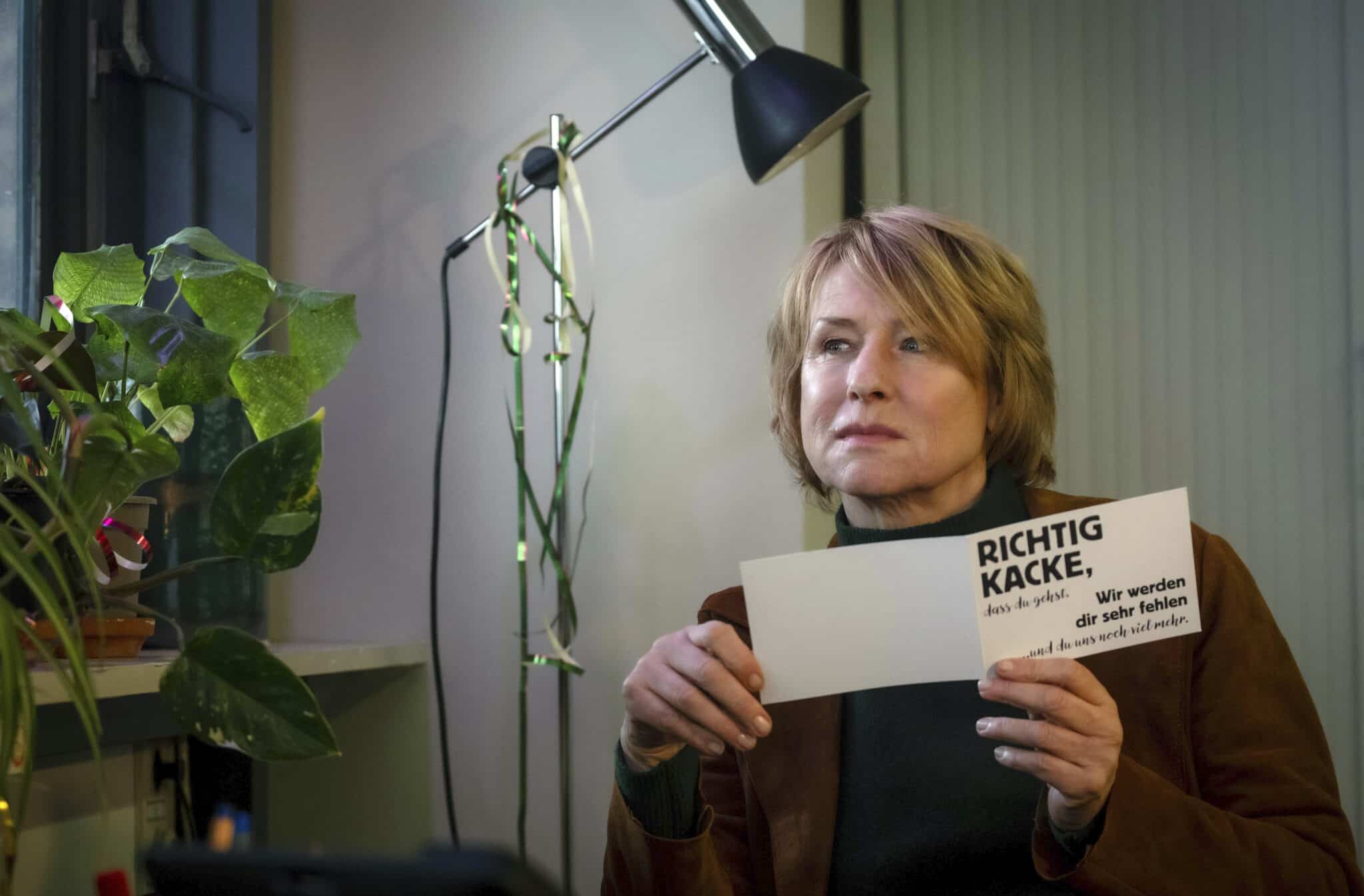 Frau mit blonden Haaren hält ein Schild mit Aufschrift in einem Raum mit Pflanzen.