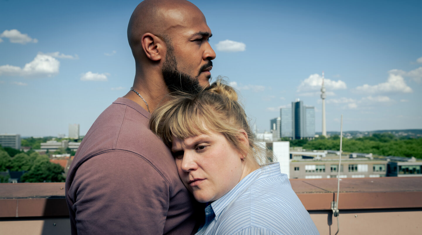 Mann und Frau stehen eng umschlungen auf einem Dach mit Stadtansicht.