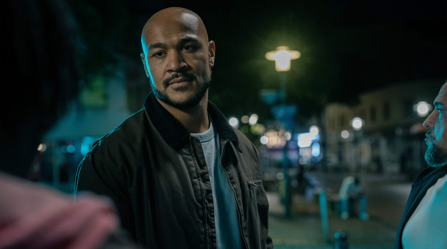 Mann mit Glatze und Jacke steht in einer nächtlichen Stadtumgebung.