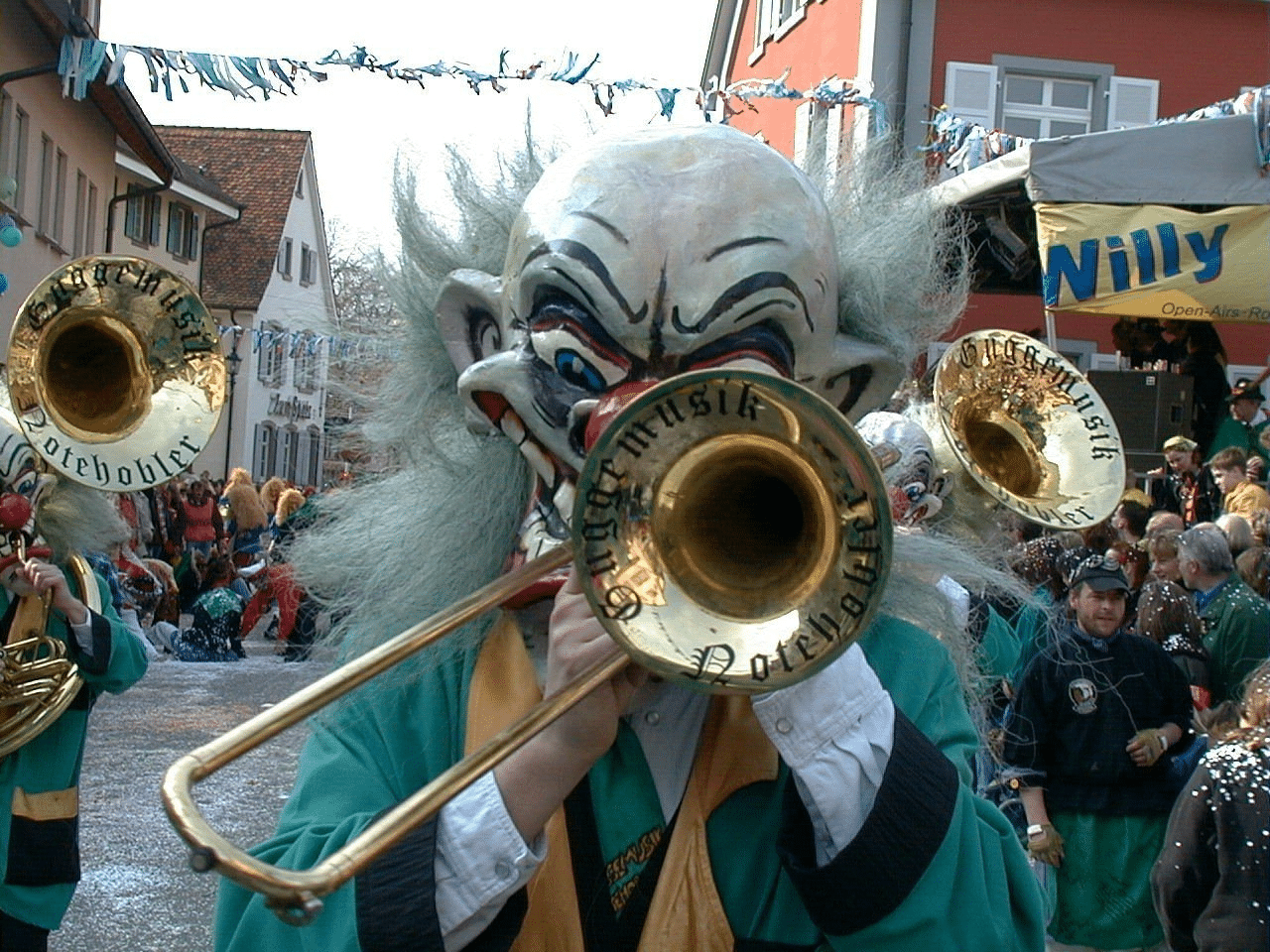 Mann mit einer großen Maske spielt Posaune bei einem Festumzug.