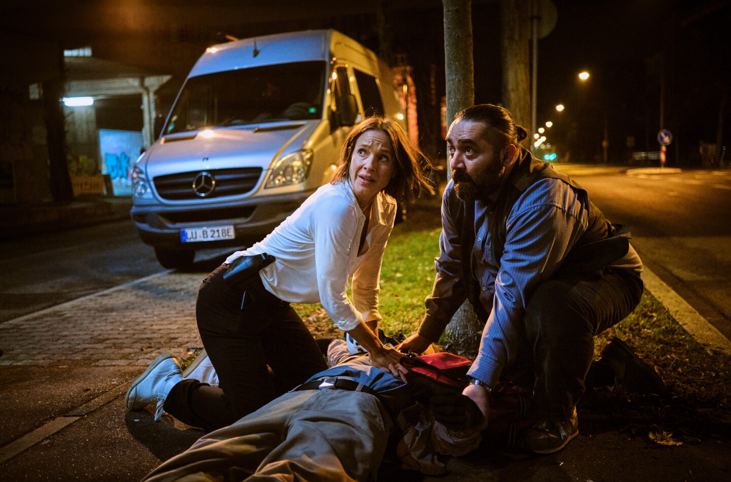 Zwei Personen helfen einem Verletzten auf der Straße bei Nacht.