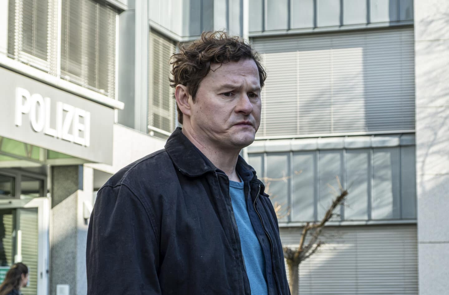 Mann mit lockigem Haar steht vor einem Polizeigebäude.