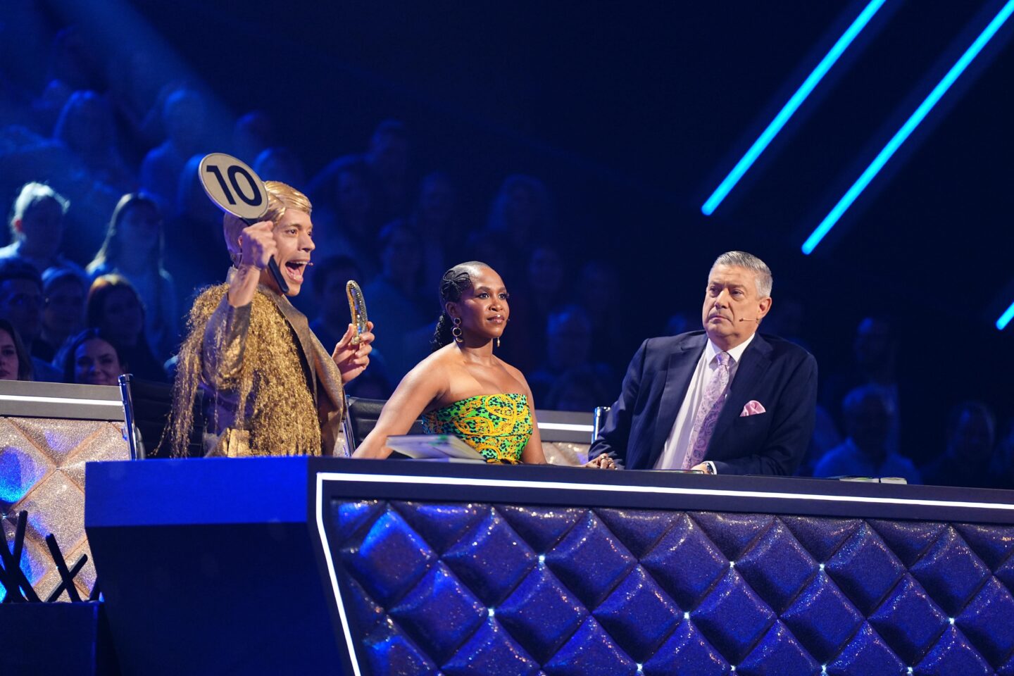 Drei Juroren in einer Show, einer hält eine Punktzahl von 10 hoch.