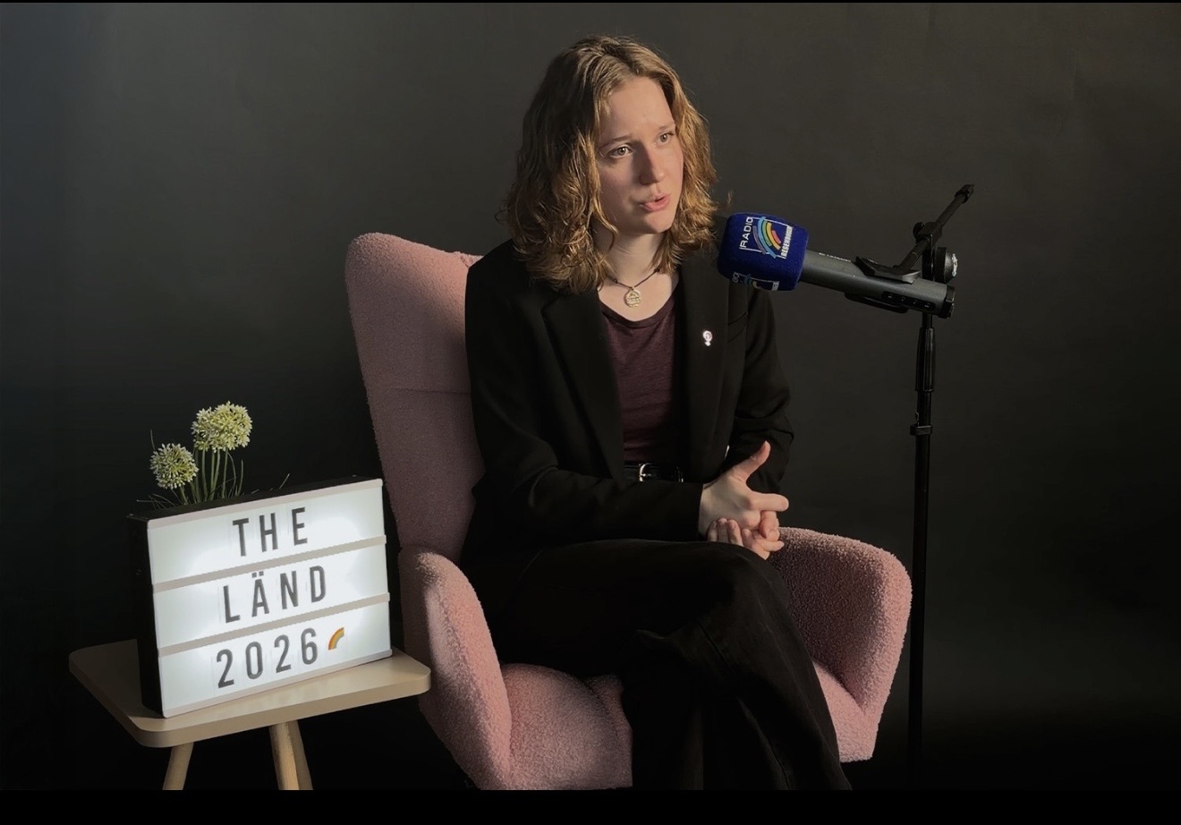 Frau im Anzug sitzt auf einem pinken Stuhl, Mikrofon und Lichtbox mit 'THE LÄND 2026' im Hintergrund.