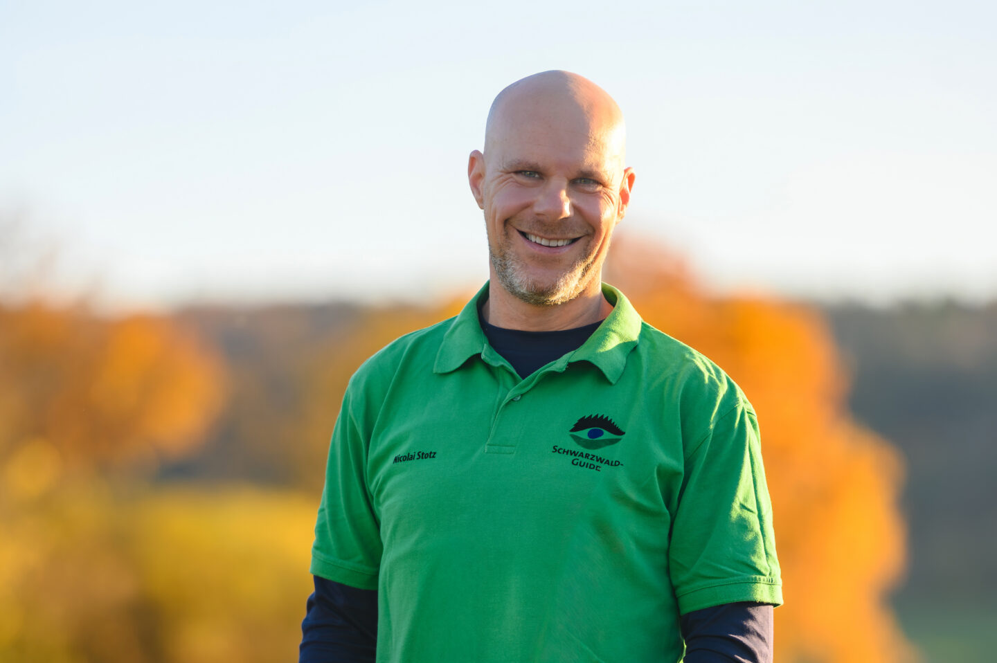 Lächelnder Mann in grünem Polohemd vor herbstlicher Landschaft.