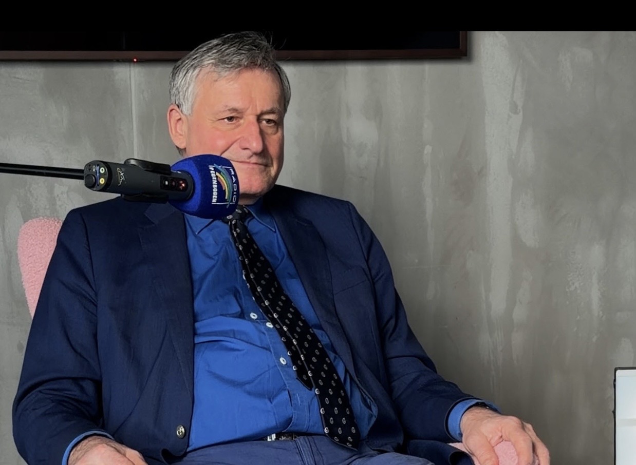 Mann im Anzug mit Mikrofon, sitzt in einem modernen Raum.