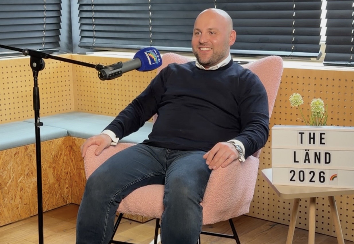 Mann sitzt in einem pinken Sessel mit Mikrofon und Schild im Hintergrund.
